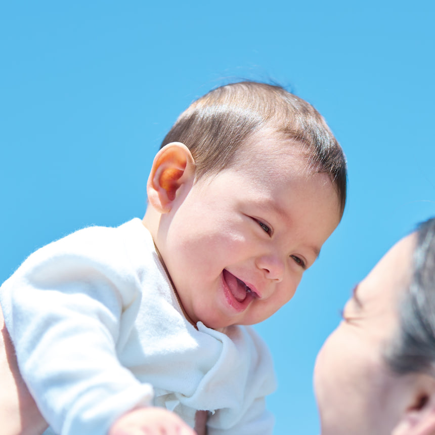 青空の下で笑顔を見せる赤ちゃん。スゴカルL コンパクトでのお出かけが楽しくなる、安心感と快適さを象徴するイメージビジュアル。