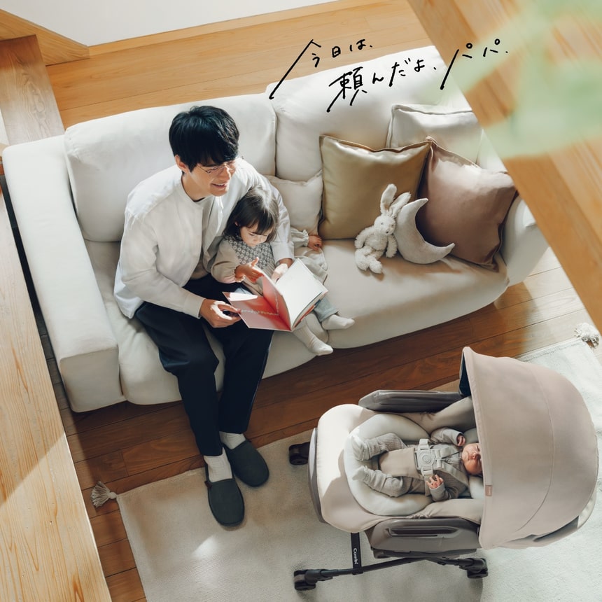 「今日は、頼んだよ、パパ」コンビ ネムリラ ベビーラックで赤ちゃんが快適にお昼寝 パパが上の子と絵本を読む穏やかなリビングシーン
